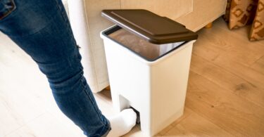 A person wearing blue jeans and white socks opens a white trashcan using their foot.