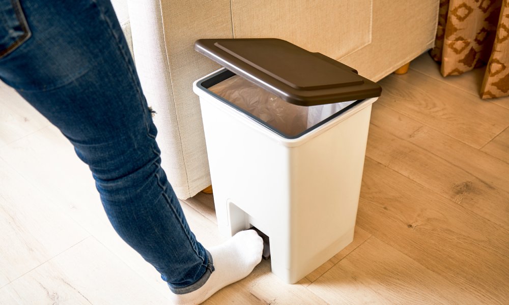 A person wearing blue jeans and white socks opens a white trashcan using their foot.