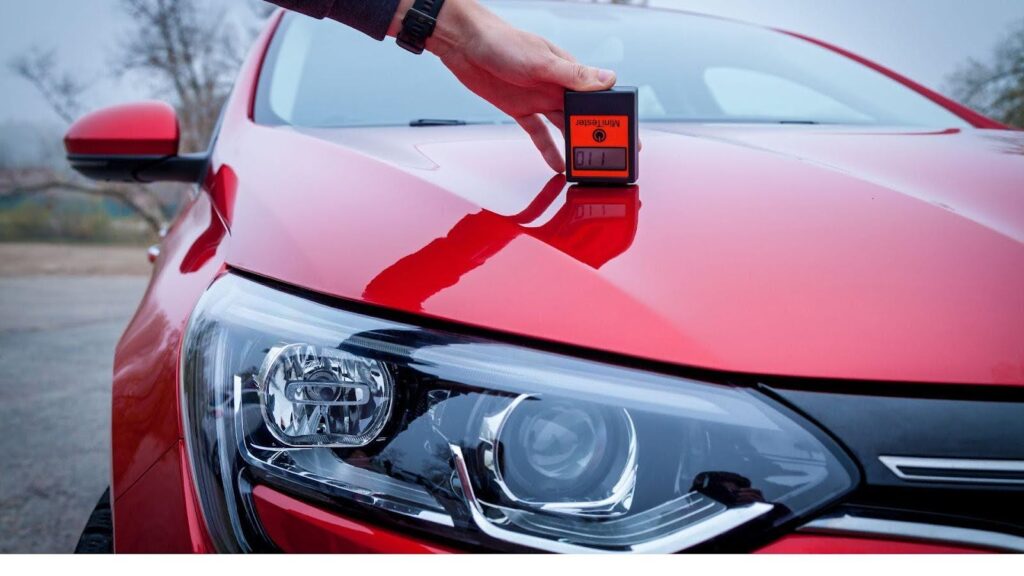 A person using a digital paint thickness gauge to measure the ceramic coating on a red car’s hood, highlighting the importance of maintaining the coating's integrity for long-term protection.