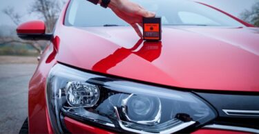 A person using a digital paint thickness gauge to measure the ceramic coating on a red car’s hood, highlighting the importance of maintaining the coating's integrity for long-term protection.