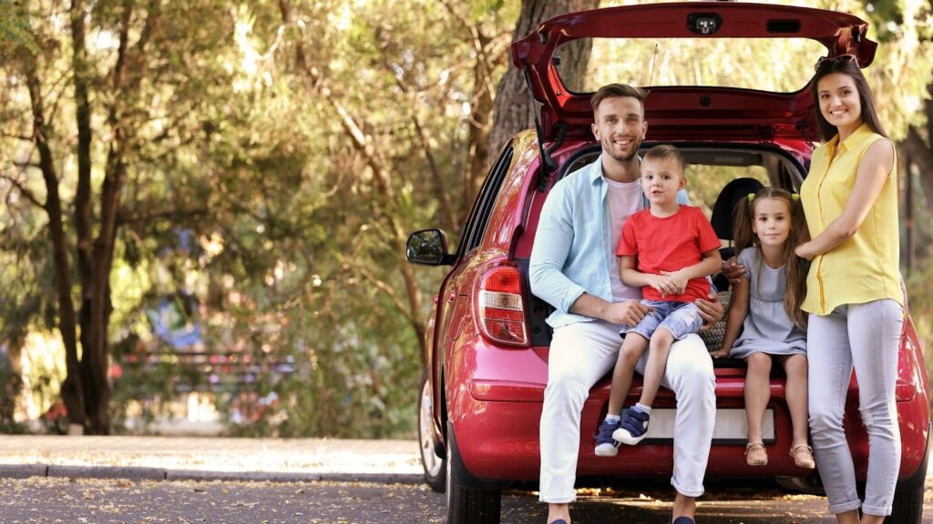 A smiling family with two young children sitting in the trunk of their red car, parked in a shaded forest area, ready for a stress-free road trip with essential vehicle fixes. 