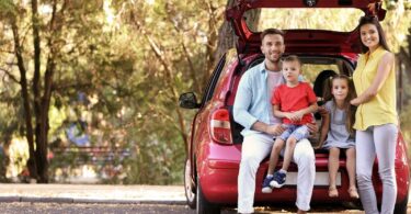 A smiling family with two young children sitting in the trunk of their red car, parked in a shaded forest area, ready for a stress-free road trip with essential vehicle fixes.