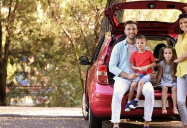 A smiling family with two young children sitting in the trunk of their red car, parked in a shaded forest area, ready for a stress-free road trip with essential vehicle fixes.