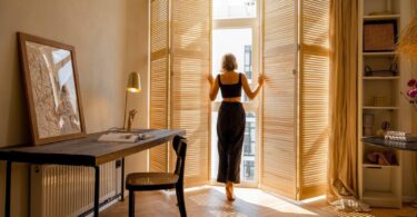 A woman opens wooden blinds in a sunlit room, set up as a cosy workspace.