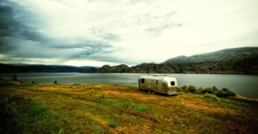 A silver airstream trailer parked next to a calm lake on a patch of orange, red, and green grass near mountains.