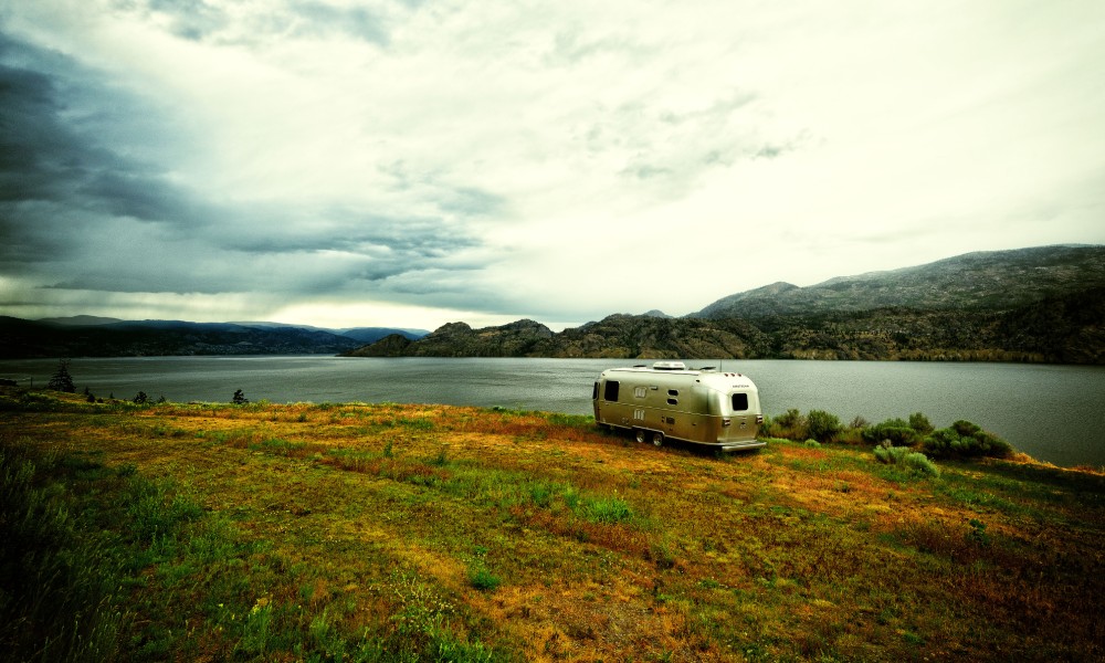 A silver airstream trailer parked next to a calm lake on a patch of orange, red, and green grass near mountains.