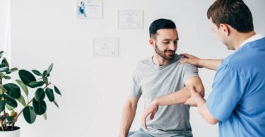 A physical therapist is with their patient, who sits on an examination bed. The therapist helps the patient with his shoulder.