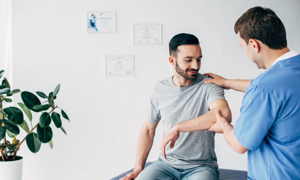 A physical therapist is with their patient, who sits on an examination bed. The therapist helps the patient with his shoulder.