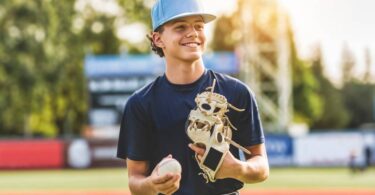 A young boy wears a blue shirt, white baseball pants, and a blue baseball hat. He holds a baseball and a baseball glove.