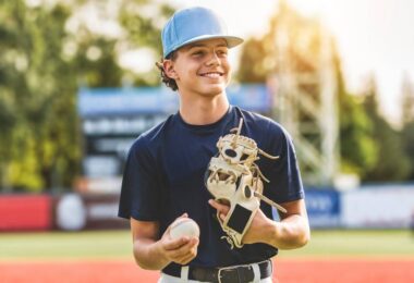 A young boy wears a blue shirt, white baseball pants, and a blue baseball hat. He holds a baseball and a baseball glove.