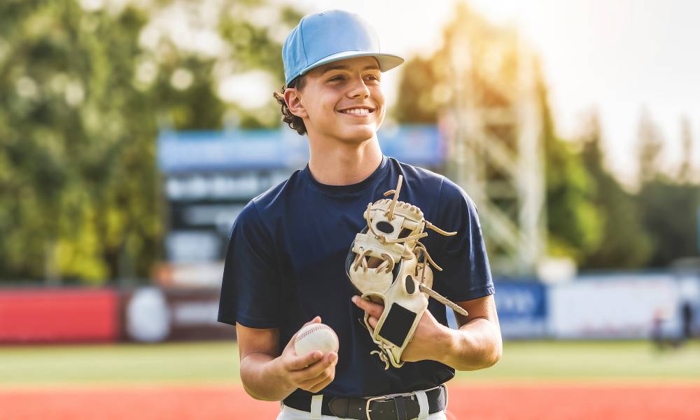 A young boy wears a blue shirt, white baseball pants, and a blue baseball hat. He holds a baseball and a baseball glove.