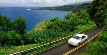 A white car drives on a two-lane stretch of the Hana Highway in Maui, Hawaii, with the ocean in the background.