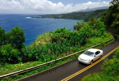 A white car drives on a two-lane stretch of the Hana Highway in Maui, Hawaii, with the ocean in the background.