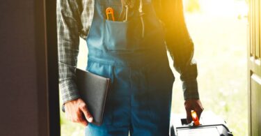 A person wearing blue overalls and a plaid shirt stands in the doorway of a home, holding a toolbox and notebook.