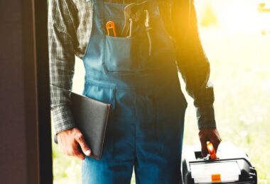 A person wearing blue overalls and a plaid shirt stands in the doorway of a home, holding a toolbox and notebook.