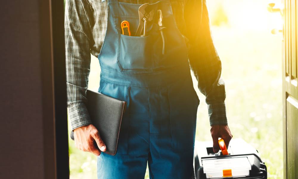 A person wearing blue overalls and a plaid shirt stands in the doorway of a home, holding a toolbox and notebook.