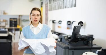 A woman in a print shop holds a crumpled and ripped piece of paper. She's looking toward the camera with a sad expression.