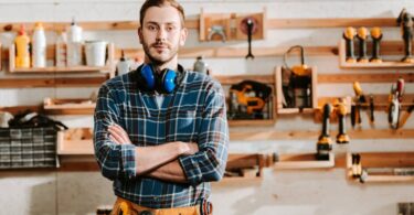 A man stands with crossed arms in a woodworking shop. He has a toolbelt and hearing protection headphones around his neck.