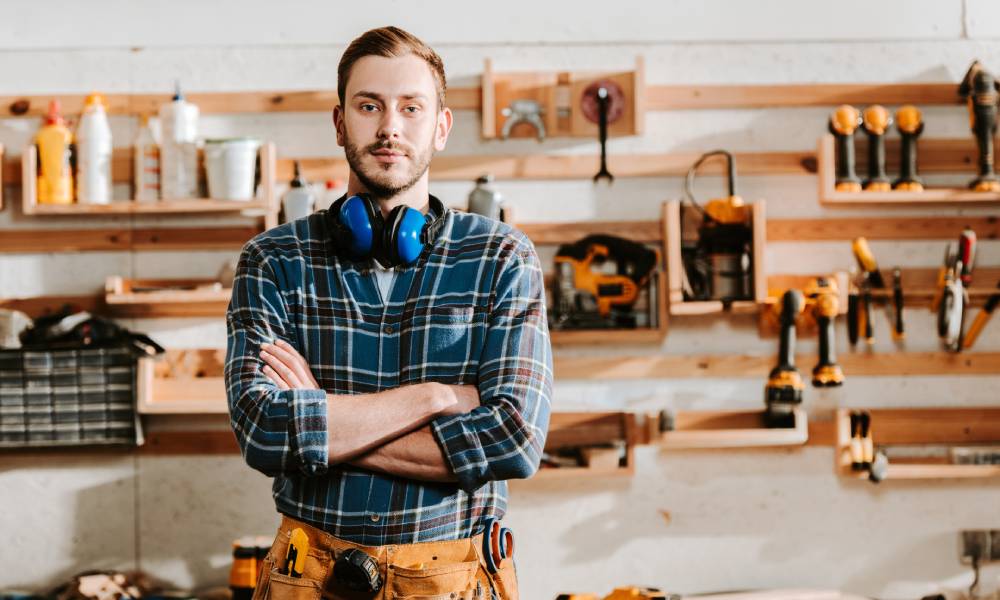 A man stands with crossed arms in a woodworking shop. He has a toolbelt and hearing protection headphones around his neck.