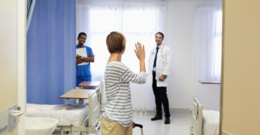 A woman making her way out of a patient hospital room with a rolling suitcase. She is turning to wave at two healthcare workers.