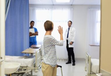 A woman making her way out of a patient hospital room with a rolling suitcase. She is turning to wave at two healthcare workers.