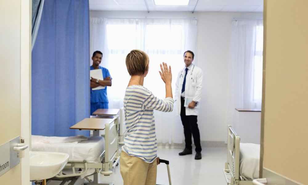 A woman making her way out of a patient hospital room with a rolling suitcase. She is turning to wave at two healthcare workers.