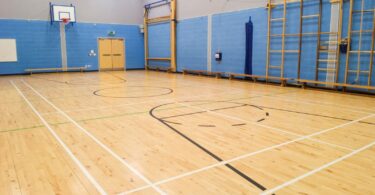 A spacious indoor gymnasium featuring a pristine wooden floor with basketball court markings, blue padded walls, and gym equipment.