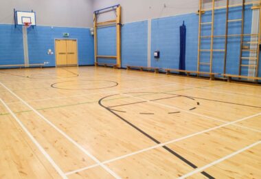 A spacious indoor gymnasium featuring a pristine wooden floor with basketball court markings, blue padded walls, and gym equipment.