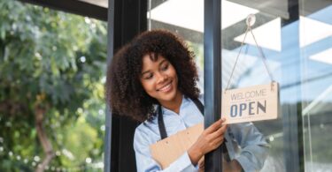 A Black small business owner peeking outside the door to flip the "open" sign. She's wearing an apron and smiling.