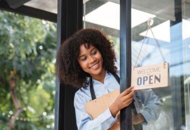 A Black small business owner peeking outside the door to flip the "open" sign. She's wearing an apron and smiling.