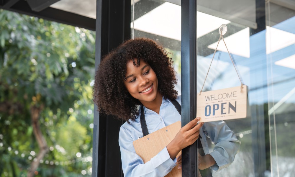 A Black small business owner peeking outside the door to flip the "open" sign. She's wearing an apron and smiling.