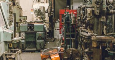 An old, cluttered, and dirty workshop with a wide variety of heavy-duty equipment sitting around. There's a fan on a table.