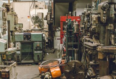 An old, cluttered, and dirty workshop with a wide variety of heavy-duty equipment sitting around. There's a fan on a table.