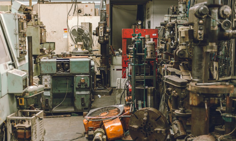 An old, cluttered, and dirty workshop with a wide variety of heavy-duty equipment sitting around. There's a fan on a table.