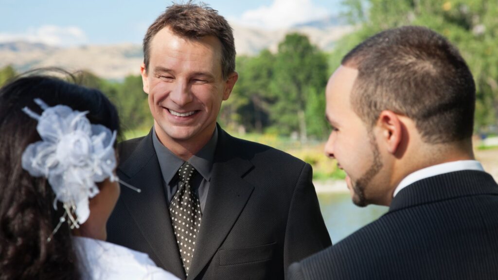 A celebrant smiling and talking with a couple during their wedding rehearsal in a park.