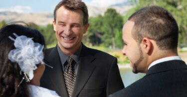 A celebrant smiling and talking with a couple during their wedding rehearsal in a park.