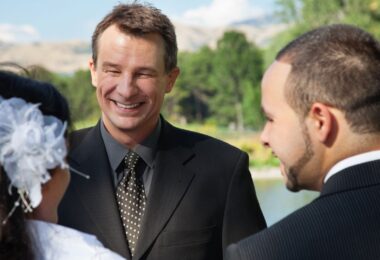 A celebrant smiling and talking with a couple during their wedding rehearsal in a park.