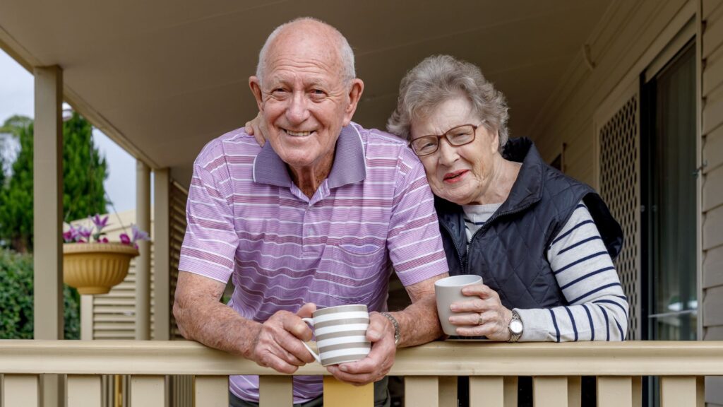 Elderly couple smiling and holding coffee mugs while leaning on a wooden porch railing.