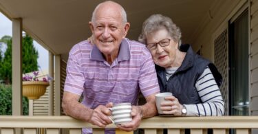 Elderly couple smiling and holding coffee mugs while leaning on a wooden porch railing.