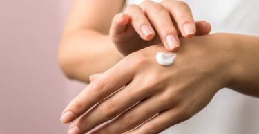 A beauty guru applying a hand cream onto their hands. The backdrop is pink and they are showing both hands.