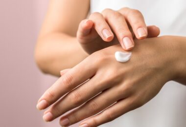 A beauty guru applying a hand cream onto their hands. The backdrop is pink and they are showing both hands.