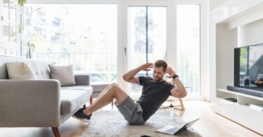 A man wearing shorts and a t-shirt sits in his living room with his feet under the couch doing a Russian twist next to a laptop.