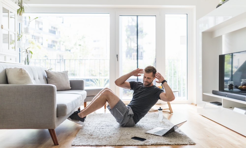 A man wearing shorts and a t-shirt sits in his living room with his feet under the couch doing a Russian twist next to a laptop.