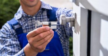 A locksmith in a blue and white plaid shirt holding a key and a cylinder lock outside a white door, exemplifying what to check before calling a 24/7 locksmith.