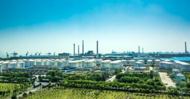 An oil refinery with white tanks for storing fuel surrounded by green trees and grass beneath a blue sky.