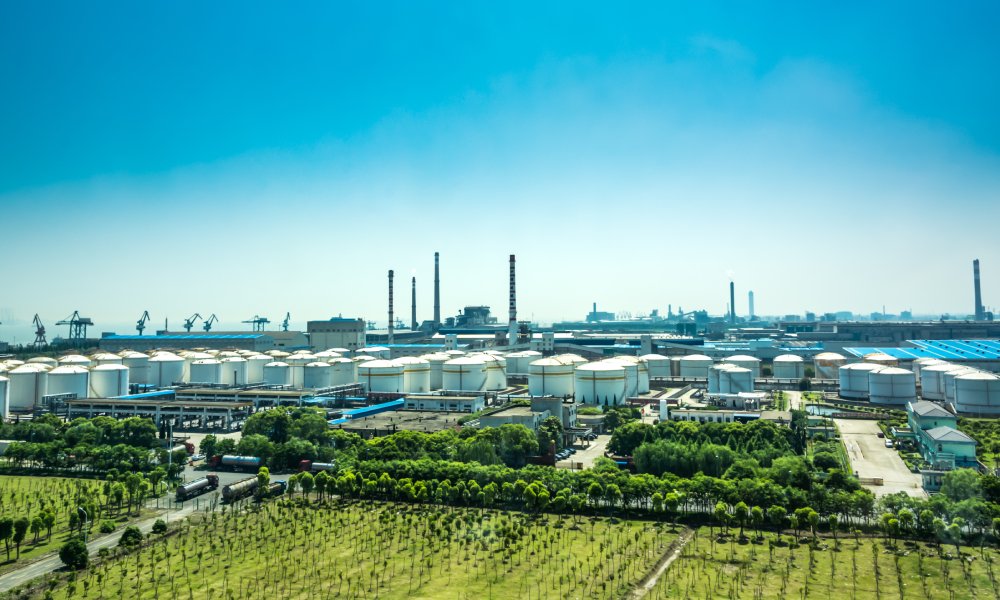 An oil refinery with white tanks for storing fuel surrounded by green trees and grass beneath a blue sky.