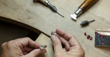 Close-up to two hands holding a small clear rock and a silver ring with multiple lapidary tools on top of a wooden table.