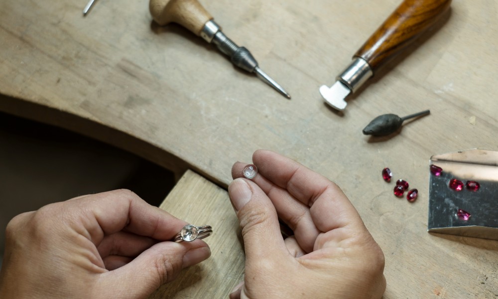 Close-up to two hands holding a small clear rock and a silver ring with multiple lapidary tools on top of a wooden table.