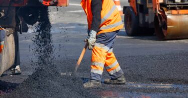 A road worker in orange overalls and protective gear shovels asphalt spilling from a machine onto a freshly paved road.