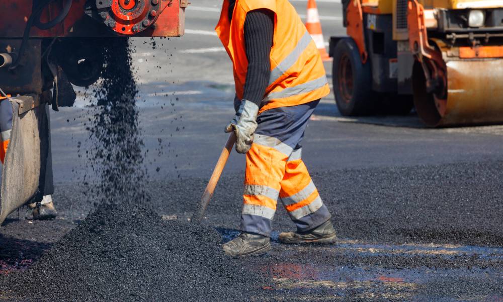 A road worker in orange overalls and protective gear shovels asphalt spilling from a machine onto a freshly paved road.